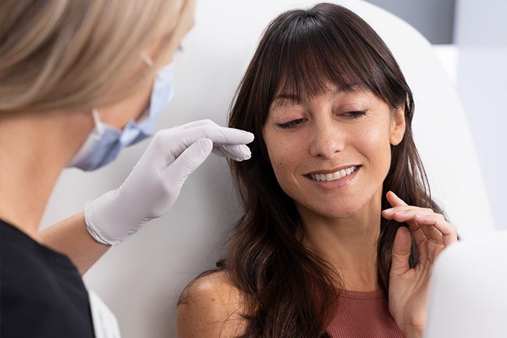 Woman wearing face mask wearing disposable gloves talking to a woman seated in examination room.