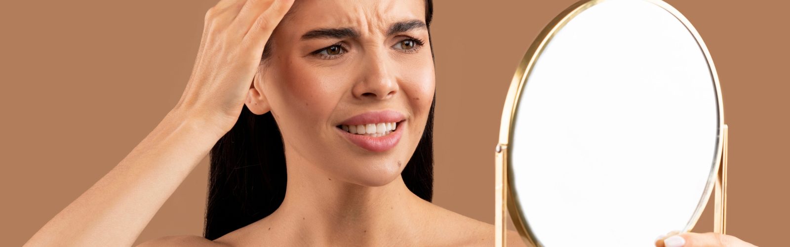 Woman holding her hand on the side of her head while staring into a mirror.