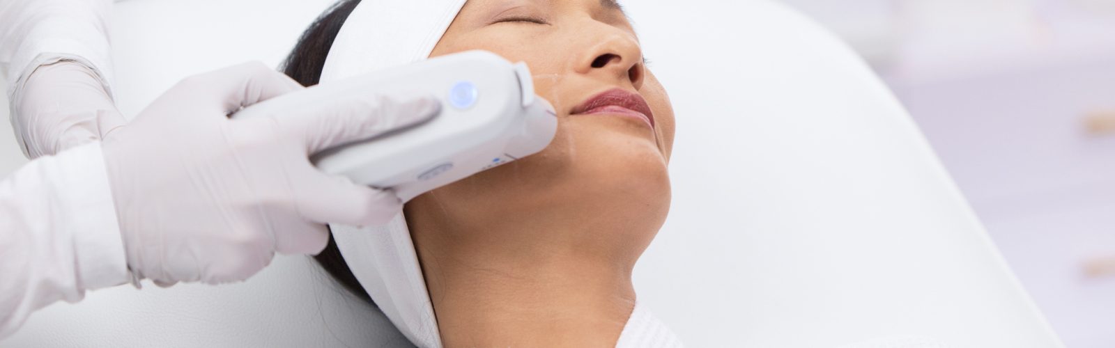 Woman with white headband laying down with her eyes closed receiving Ultherapy on her face.