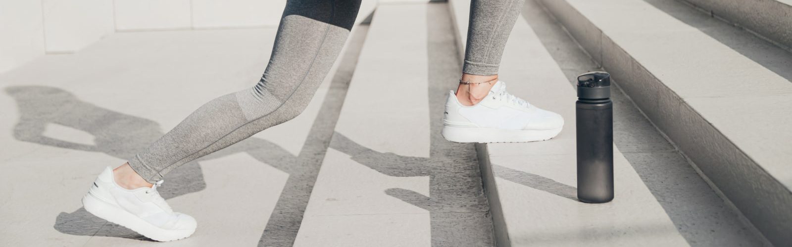 Closeup of person wearing grey leggings and white tennis shoes walking up stairs with a water bottle.