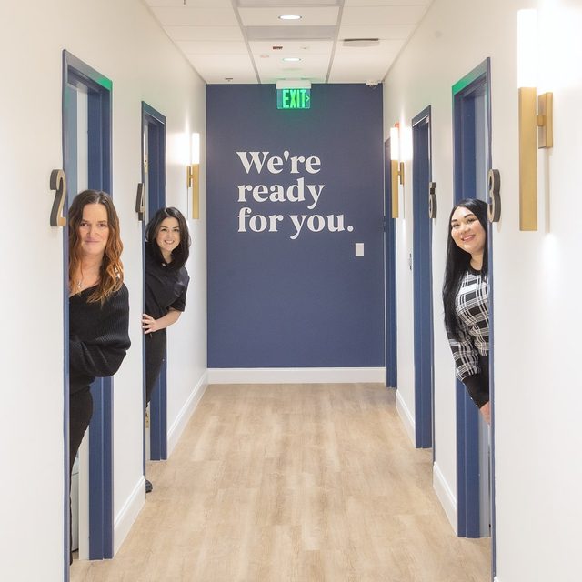 Three women in the doorway of a clinic with a sign on the wall saying, "We're ready for you."