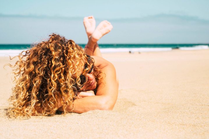 Adult woman with curly hair laying on the sand near a beach.