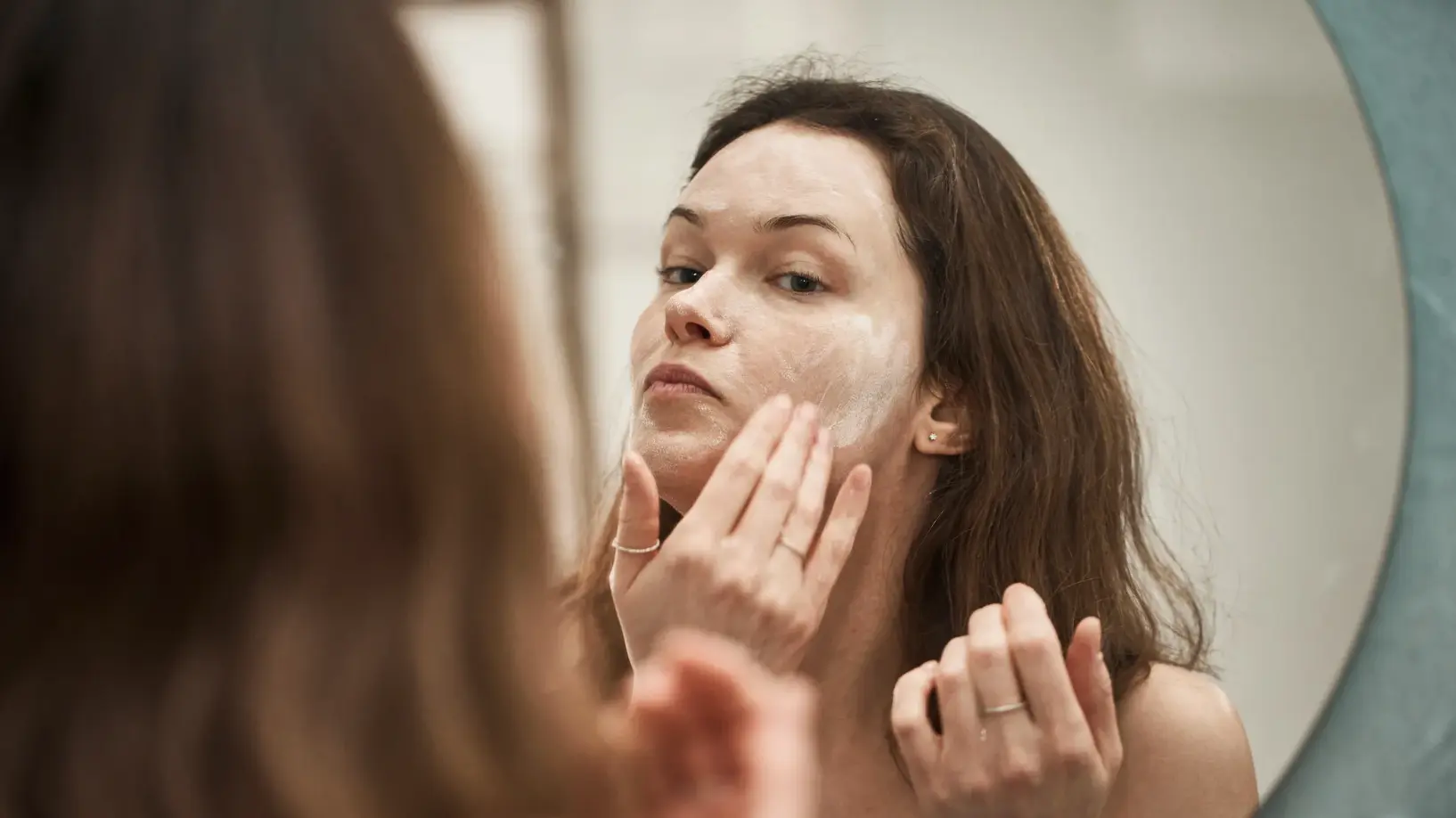 Young woman applying face cream in mirror